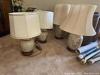 Four ceramic floral table lamps of varying heights on carpeted floor, with rolled posters nearby.