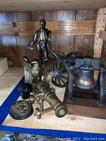 Photo showing arrangement of metal art collection including standing figure, brass eagle, brass cannon, black metal skull, paperweight, and Liberty Bell replica.