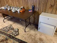 Metal folding table with dark brown top and black metal legs, placed against wood panel wall with carpeted floor; shows some scratches on table surface.