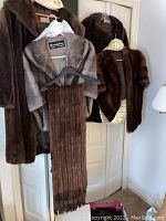 Four fur items hanging on wall-mounted hooks: long fox fur coat, mink fur jacket with muff, dark brown fur jacket, and brown fur stole.