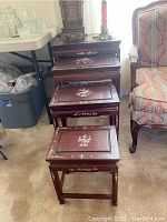 Four wooden nesting tables stacked from largest to smallest. Tables have decorative white inlay floral patterns with geometric border designs. Visible surface scratches and wear.