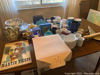 Wide view showing collection of ceramic mugs, plates, bowls, a wooden desk organizer, and model airplane on a table.