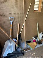 Photo showing metal shovel, garden rake, leaf rake, and broom against a pegboard wall