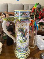 Image showing hand-painted ceramic tankard vase with rooster design, figurines, glass jar with colorful wooden bird picks in background.