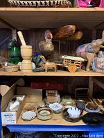 Shelf display of wooden South American art including bird sculpture, wooden mortar and pestle, miniature wagon, and ox figure, along with ashtrays and small ceramic items below the shelf.