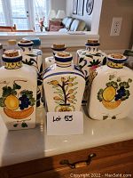 Front view of six assorted shaped ceramic jars with various hand-painted fruit motifs and cork stoppers arranged on counter.