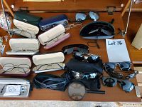 Top view showing multiple eyeglasses and sunglasses on wooden surface, some in cases, some out, plus magnifying glass and various cases.