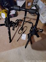 Front view of black metal car bike rack with straps and foam padding on arms, photographed indoors on carpet.