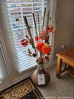 Full view of tall clay vase with flower arrangement near window and table.