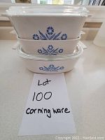 Five white ceramic CorningWare casserole dishes with blue cornflower patterns stacked in three rows. Three dishes have clear glass lids.