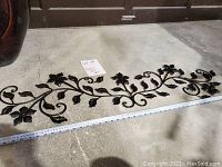 Full view of the black metal floral wall art laid on the floor with a measuring tape showing approximately 60 inches length.