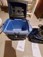 Blue mini cooler with its lid open, showing interior, next to cooler bag and accessories on floor.