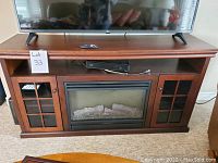 Front view of the Muskoka fireplace TV stand with a flat-screen TV on top, remote on shelf, and fireplace insert visible in the middle along with side shelving and glass grid doors.