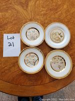 Four chokin decorative plates arranged on a wooden table, each with a different nature-themed gold accented design in the center and a gold outer rim. Plates have metal hangers attached for wall mounting.