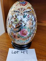 Photo shows a large, oval pheasant egg with intricate colorful floral and bird designs, resting on a black wooden stand. The egg is glossy with detailed paintwork.