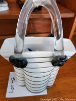 White glass vase with horizontal black stripes and clear arched handles shown from a front angle with wooden background and other items visible.