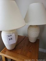 Pair of beige ceramic table lamps with textured base and white tapered lampshades, placed on a wooden side table.