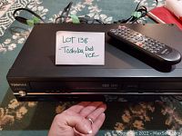 Toshiba black DVD and VHS combo player showing front panel with remote on top and a hand for scale.