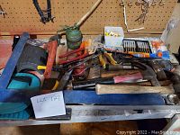 Wide view showing assortment of hand tools including clamps, oil can, scissors, drill bits, and brush on workbench surface.