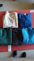 Photo showing five sweaters and vests laid out on a red table: one light grey cardigan and four V-neck sweaters in shades of blue, green and charcoal.