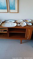 Photo of assorted china plates and platters displayed on wooden cabinet showing size and variety.