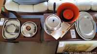 Overview of the full set showing fondue pot, enamel bowls in orange and blue, metal serving trays and dishes arranged on tables and chair.