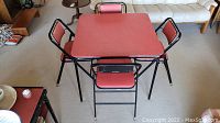 Square metal folding card table with red laminate top and black frame surrounded by 4 matching folding chairs with red padded seats and backs.