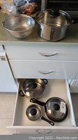 Four stainless steel pots with lids inside a drawer, a frying pan lid visible, and a metal mesh strainer on countertop.