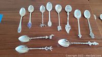 Full set of 12 silver collector spoons displayed on wood surface showing various decorative designs and shapes.