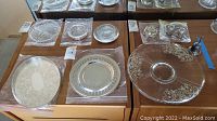 Various silver plated and glass serving dishes arranged on a wooden surface, showing items in the lot including plates, trays, and a glass cake plate.