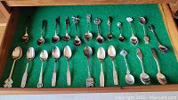 View showing 20 assorted silver plated collector spoons placed on green felt, displaying a variety of handle designs and shapes.