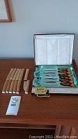 Wide shot showing both sets of steak knives: one set with wooden handles laid out on table and another set of six in original presentation box with light blue satin lining and cream interior.