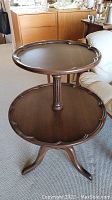 Side and angled views of a two tiered wood table showing scalloped edges on the round tabletops, turned central column, and three curved legs.