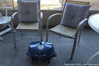 Two metal patio chairs with beige seats and gray cushions, with blue Uniflame tabletop grill in front on concrete floor.