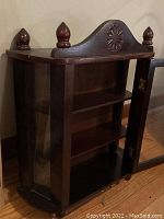 Side view of antique wooden display shelf showing door with glass panel, two shelves inside, carved top with floral design and finials