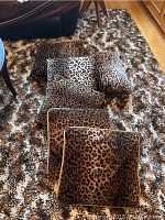 Six leopard print velvet cushions arranged on matching leopard print rug/floor covering. Cushions vary in brown, tan, and cream leopard patterns with some edged in black piping.