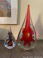 Two glass art pieces, tallest 11 inches and shortest 6 inches, both teardrop-shaped with colored internal design elements, set against a wall with framed artwork.