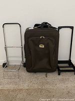 Front view of brown soft-sided upright suitcase with extendible handle and two different luggage carts placed on either side against a wall.
