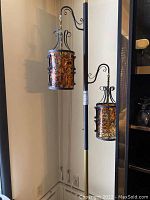 Full view of the black metal tension pole floor lamp with two hanging lanterns showing the amber and brown mottled glass panels and wrought iron scrollwork.