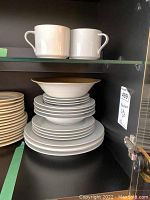 View of stacked plates, shallow bowls with gold trim, and two white porcelain cups on glass shelf in cabinet