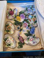 Twelve miniature floral porcelain placeholders and ashtrays displayed in blue box with tissue paper cushioning. Colorful flowers and green leaves attached to white porcelain bases with gold trim.