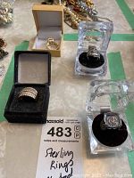 Four rings with three marked sterling silver, displayed in various jewelry boxes and clear display cases, on a table with a MaxSold lot note.