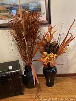 Two black ceramic vases with dried floral arrangements, one with orange autumn leaves and another with tall brown grass-like materials.