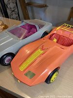 Photo showing two vintage Barbie Corvette cars: one silver and one orange, displaying their condition and color details.