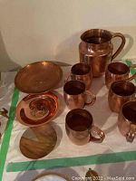 Photo showing 6 copper Moscow Mule mugs arranged near 3 decorative copper plates, with one plate on a stand.