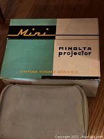 Original Minolta projector box with green, black, and cream coloring, labeled Mini Minolta projector, alongside fabric carrying case.
