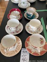 Six vintage tea cups and saucers arranged on black table, showing variety of patterns and colors including reds, turquoise, and citrine tones.