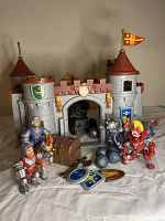 Frontal view of vintage gray plastic toy castle with two conical red-topped towers featuring heraldic flag designs. Five plastic figurines in knight armor and a horse are positioned in front of the castle gate. A treasure chest and several cannonballs are placed alongside figurines.