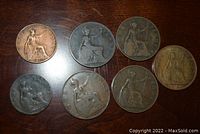 Seven British coins arranged on a wooden surface showing seated Britannia design, including pennies and half pennies, in various conditions.