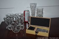 Clear glassware items including stemmed glasses, bowls, and vases with a wooden box open showing cheese knives beside a soda can for scale.
